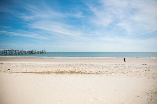 A beach with a pier extending into the ocean, a person walking on the sand, and a clear sky.