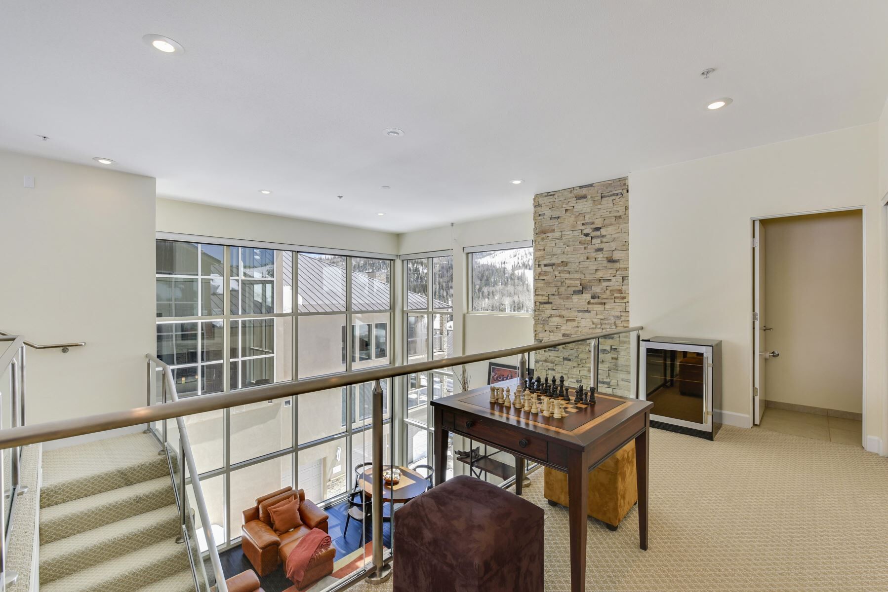 A spacious living area with a large window, a stone accent wall, a wooden table with chess pieces, a brown leather sofa, and a staircase with metal railings.