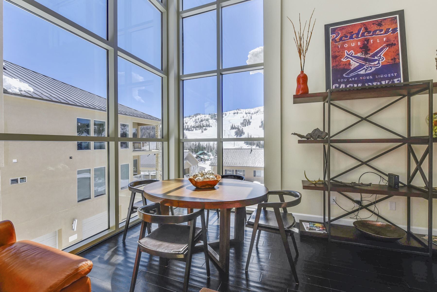 A cozy room with a large window overlooking a snowy landscape. There's a round wooden table with chairs, a small shelf with decorative items, and a framed poster on the wall.