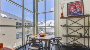 Une pièce chaleureuse avec une grande fenêtre donnant sur un paysage enneigé. On y trouve une table ronde en bois, entourée de chaises, une petite étagère avec des objets décoratifs et une affiche encadrée accrochée au mur.