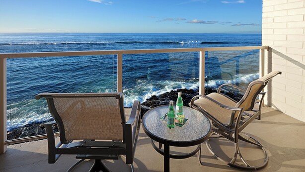 Ein Balkon mit zwei Stühlen und einem Tisch, mit Blick auf das Meer, wo Wellen an ein weißes Gebäude schlagen.