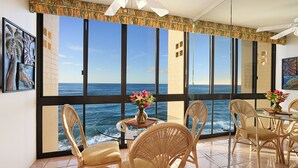 Ein Zimmer mit großem Fenster und Meerblick, Korbstühle, ein Glastisch mit Blumenvase und Deckenventilator.