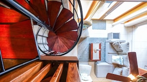 Una escalera de caracol con escalones de madera y barandillas metálicas, que conduce a una habitación con un sofá, cojines y una mesita.