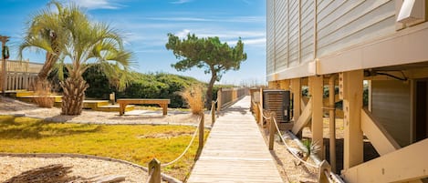 Un sendero de madera que lleva a un edificio de techo inclinado, rodeado de palmeras y un área con césped.