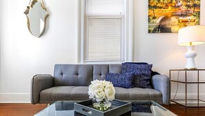 Una sala de estar moderna con un sofá gris, una mesa de centro de vidrio con un jarrón de flores blancas, una mesita de noche con una lámpara y un cuadro en la pared.