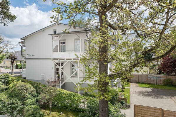 A two-story white villa with a balcony, surrounded by lush greenery and a paved driveway.