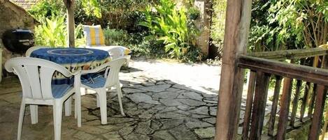 A stone patio with white plastic chairs and a table, surrounded by lush greenery and a wooden fence.