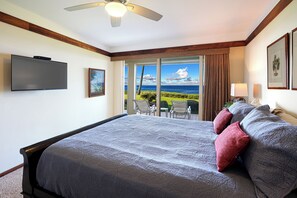 Ein Hotelzimmer mit einem großen Bett, einem Flachbildfernseher, einem Deckenventilator, einer Schiebetür zu einem Balkon mit Meerblick und einem kleinen Tisch mit Stühlen.