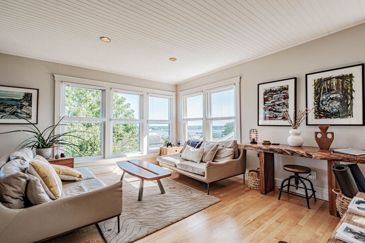 A spacious living room with a large window, a wooden coffee table, a sofa, and a dining area with a wooden table and bar stool.