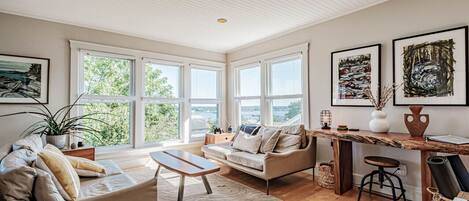 A spacious living room with a large window, a wooden coffee table, a sofa, and a dining area with a wooden table and bar stool.