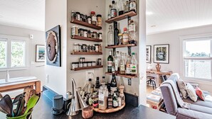 Una cocina con estantes de madera que contienen varias botellas y frascos, un comedor con mesa y sillas, y una sala de estar con sofá y ventana.