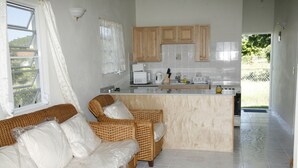 Eine Küche mit Holdelementen, einer weißen Arbeitsplatte und einer eingebauten Kochinsel. Im Wohnbereich befindet sich eine Rattan-Couch mit weißen Kissen.