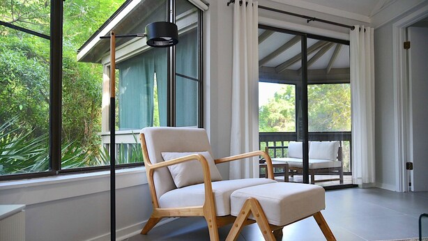 A modern room with a large window, a wooden chair, a floor lamp, and a view of greenery outside. - Newly renovated cottage in Sea Pines. Private setting with a short walk to beach (Hilton Head Island)