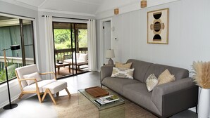 A modern living room with a grey sofa, a wooden chair, a glass coffee table, and a view of greenery through large windows. - Newly renovated cottage in Sea Pines. Private setting with a short walk to beach (Hilton Head Island)