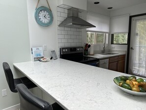 A modern kitchen with a marble countertop, a stove, oven, and a sink. There is a bowl of fruit on the counter and a clock on the wall. - Oceanfront Seaside Vacay with stunning views at Inn of the Sea,Pool/Hot Tub/Dock (Ladysmith)