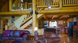 Interior espacioso de una cabaña de troncos con sala de estar que incluye sofás de cuero y un comedor. Una escalera conduce a un nivel superior.