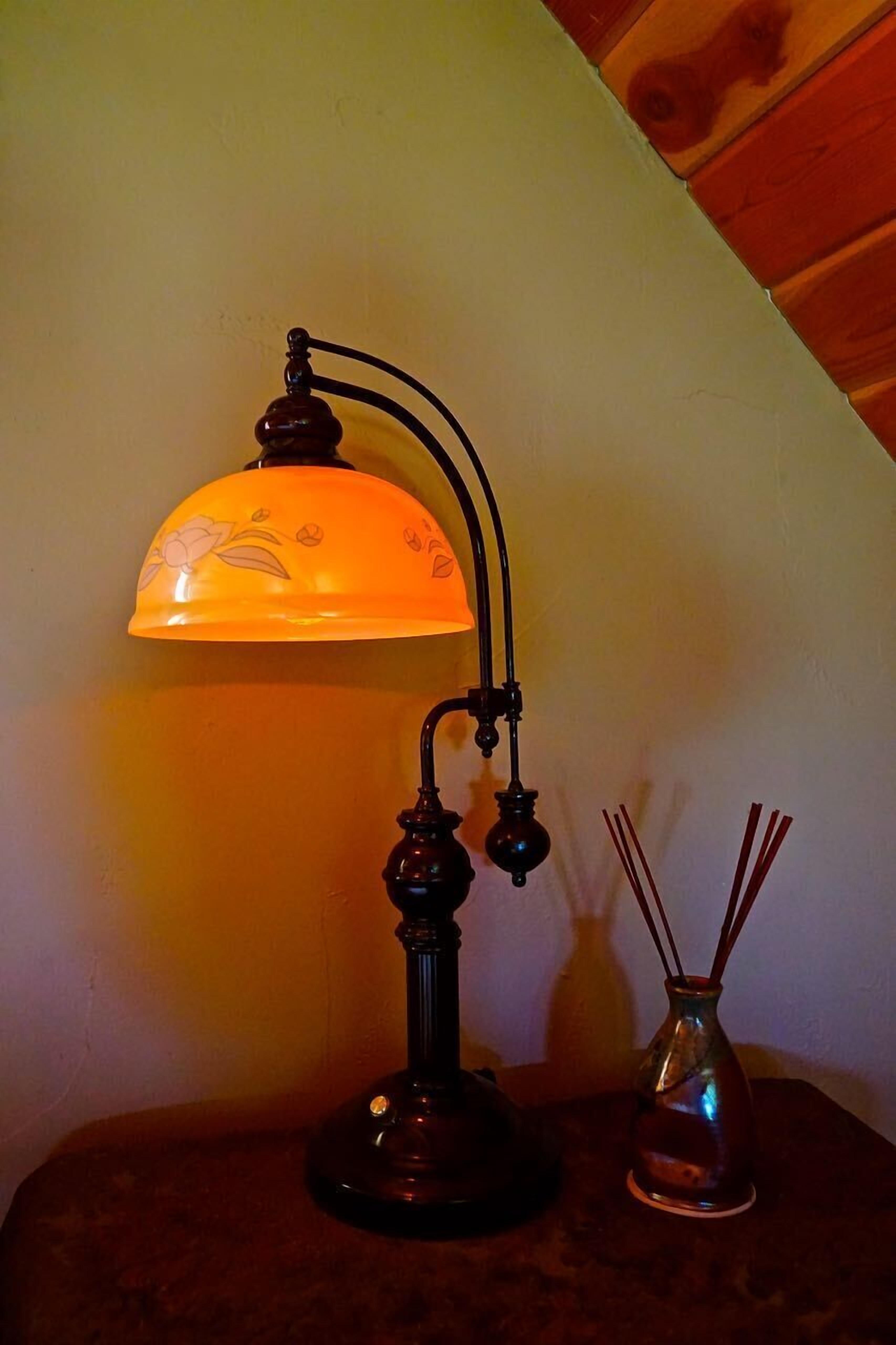 Une lampe de table avec un bras incurvé et un abat-jour en forme de dôme, un petit vase contenant des tiges de roseau et une table en bois adossée à un mur.