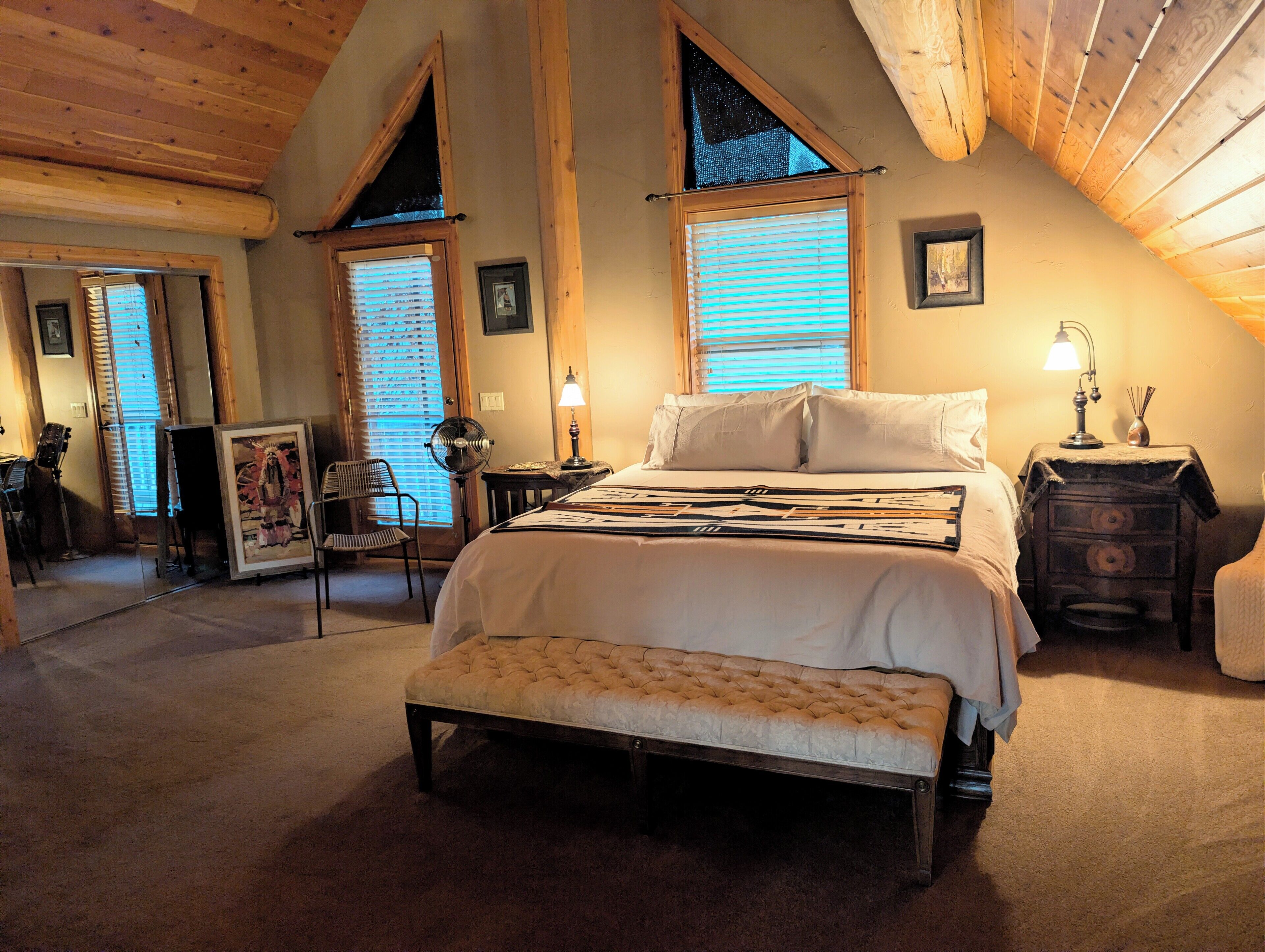 A cozy bedroom with a bed, a bench, a desk, and a chair. There are two windows with blinds, a lamp, and a picture frame on the wall.