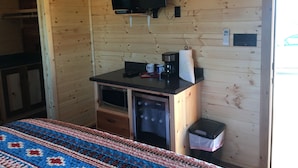 Puinen mökkimainen huone, jossa on sänky, seinälle asennettu televisio, pieni pöytä, jossa on kahvinkeitin, ja roskakori.