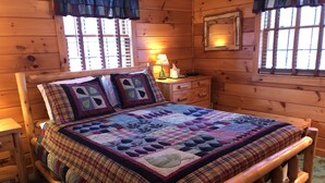 Un dormitorio de cabaña de madera con cama, mesita de noche, lámpara y ventana con contraventanas.