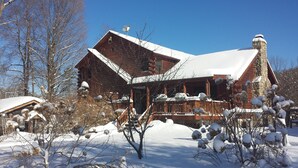 Una cabaña de troncos con chimenea de piedra, rodeada de árboles cubiertos de nieve y una terraza de madera.