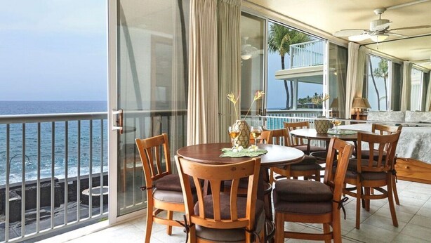 A balcony with a view of the ocean, furnished with wooden chairs and a table, and a ceiling fan.