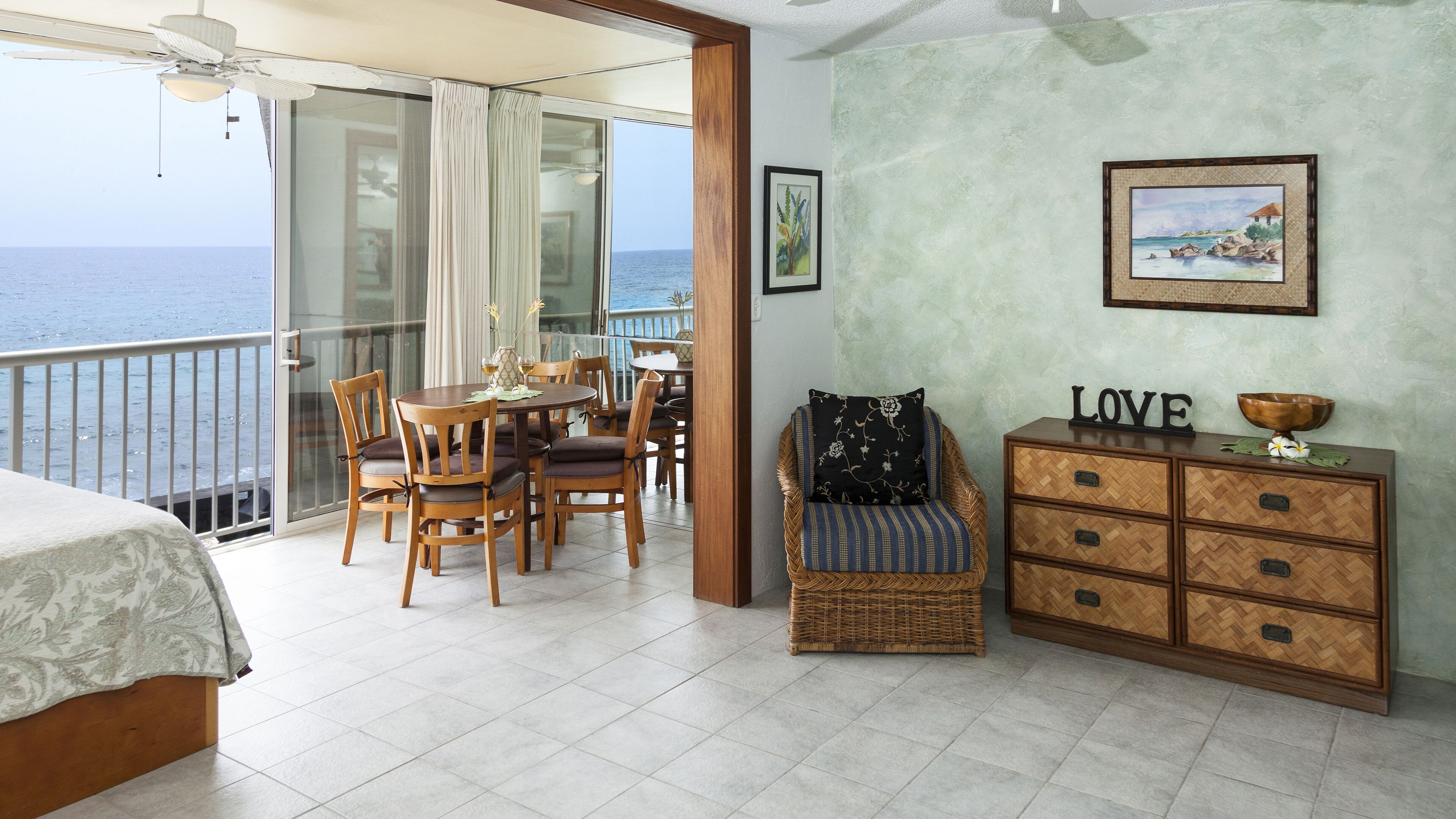A balcony with a view of the sea, a dining area with wooden chairs and a table, a wicker chair, and a wooden cabinet with drawers.