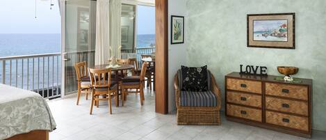 A balcony with a view of the sea, a dining area with wooden chairs and a table, a wicker chair, and a wooden cabinet with drawers.