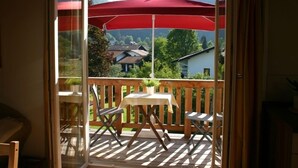Un balcón con mesa y sillas, una sombrilla roja y vistas a casas y montañas.
