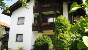 Um edifício de dois andares com varanda decorada com flores vermelhas, cercado por vegetação.