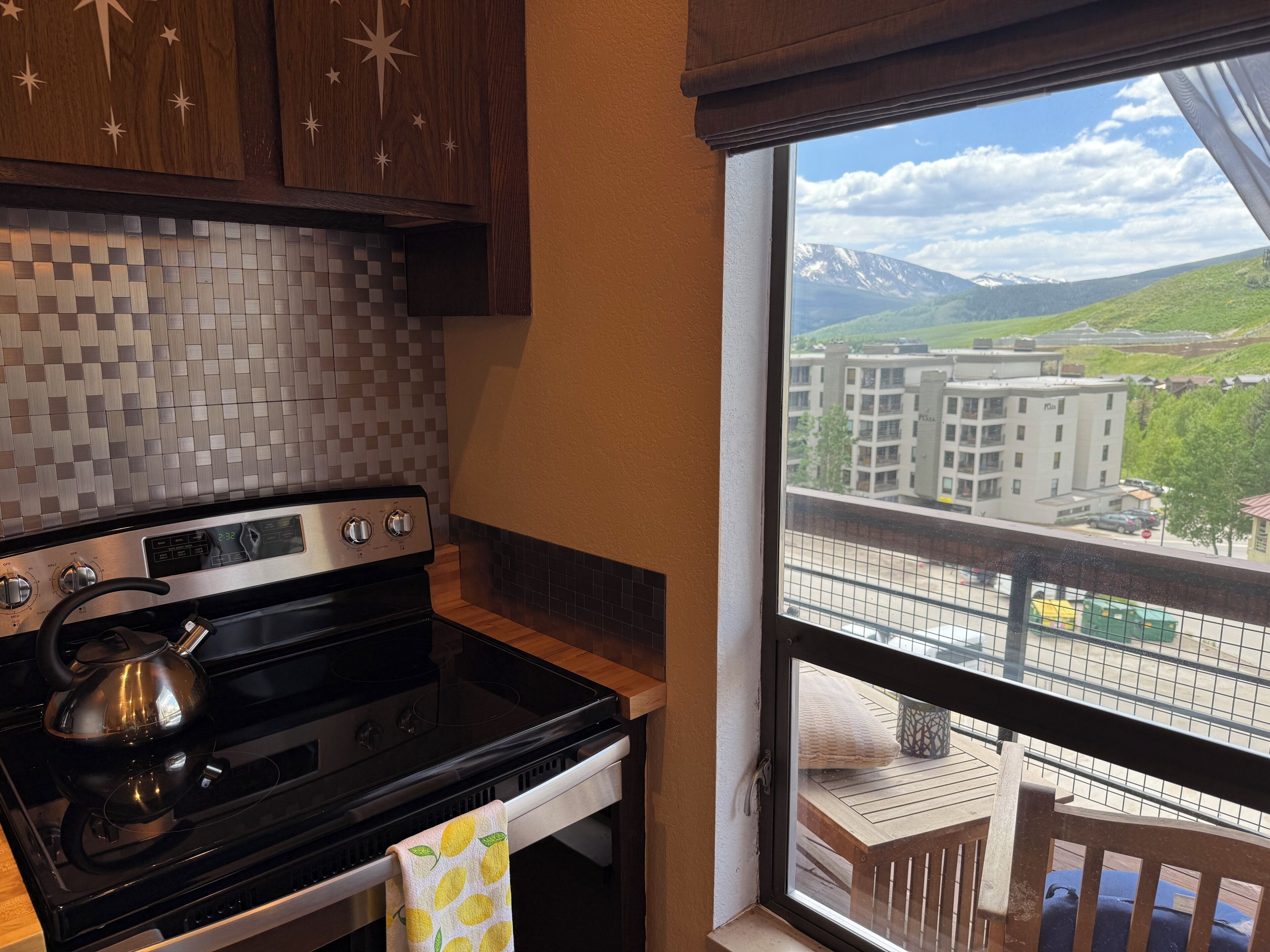 A kitchen with a stove, a kettle, and a window with a view of buildings and mountains.
