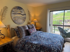Een slaapkamer met een groot bed, twee nachtkastjes met lampen, een schilderij aan de muur en uitzicht op een groen landschap vanaf een balkon.