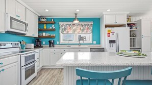 Una cocina con armarios blancos, una isla central, un microondas, una cafetera y una ventana con cortina estampada.
