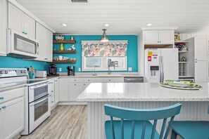 Una cocina con armarios blancos, una isla central, un microondas, una cafetera y una ventana con cortina estampada.