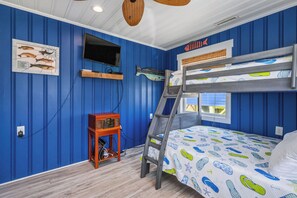 A room with blue walls, a bunk bed with a colorful mattress, a mounted TV, and a shelf with books and a fish illustration. - Moon Struck: Oceanfront Home with Bold, Coastal Flair (Oak Island)