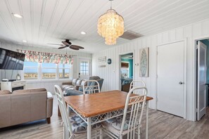 A room with a wooden dining table, white chairs, a ceiling fan, and a large window with a view of the beach. - Moon Struck: Oceanfront Home with Bold, Coastal Flair (Oak Island)