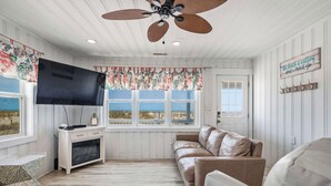 Una habitación con ventilador de techo, televisor de pantalla plana, sofá, banca y ventana con vista.