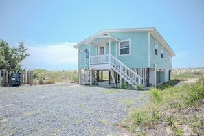 Une maison de plage à deux étages, dotée d’un porche et d’un escalier menant à l’entrée.