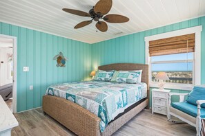 Une chambre à coucher comprenant un lit, une chaise en osier, une table de chevet, un ventilateur de plafond et une fenêtre donnant sur un paysage agréable.