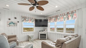 A room with a ceiling fan, a flat-screen TV, a sofa, a chair, and a window with a view of the beach. - Moon Struck: Oceanfront Home with Bold, Coastal Flair (Oak Island)