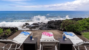 Un salón junto a la playa con tres tumbonas, cada una con una toalla y una bebida, con vistas al océano y olas rompiendo contra rocas oscuras.