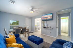 Ein modernes Wohnzimmer mit einem blauen Sofa, einem Couchtisch und einem Deckenventilator. Es gibt einen Essbereich mit Tisch und Stühlen, und im Hintergrund eine Küche.