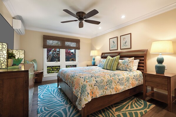 A bedroom with a bed, a ceiling fan, a window with blinds, a bedside table with a lamp, and a nightstand with a lamp. - Hale Honu | Private Oceanfront Retreat with Turtle & Bay Views (Koloa)