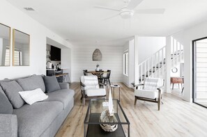 Ein modernes Wohnzimmer mit einem grauen Sofa, einem Glastisch und einem Essbereich mit einem weißen Tisch und Stühlen.