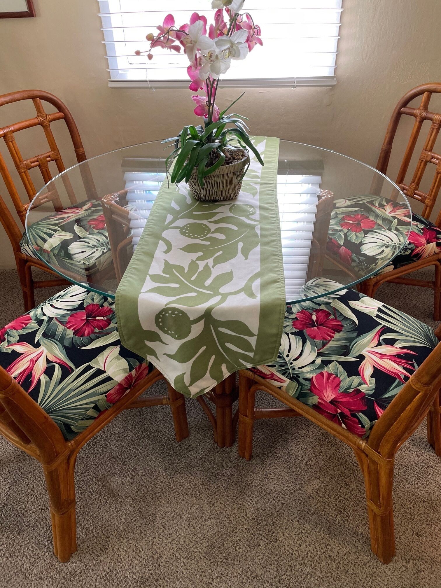 Una mesa redonda de vidrio con camino de mesa floral, rodeada por sillas de mimbre con cojines estampados.