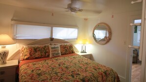 A bedroom with a bed, a ceiling fan, a nightstand with a lamp, a window with blinds, and a mirror on the wall.