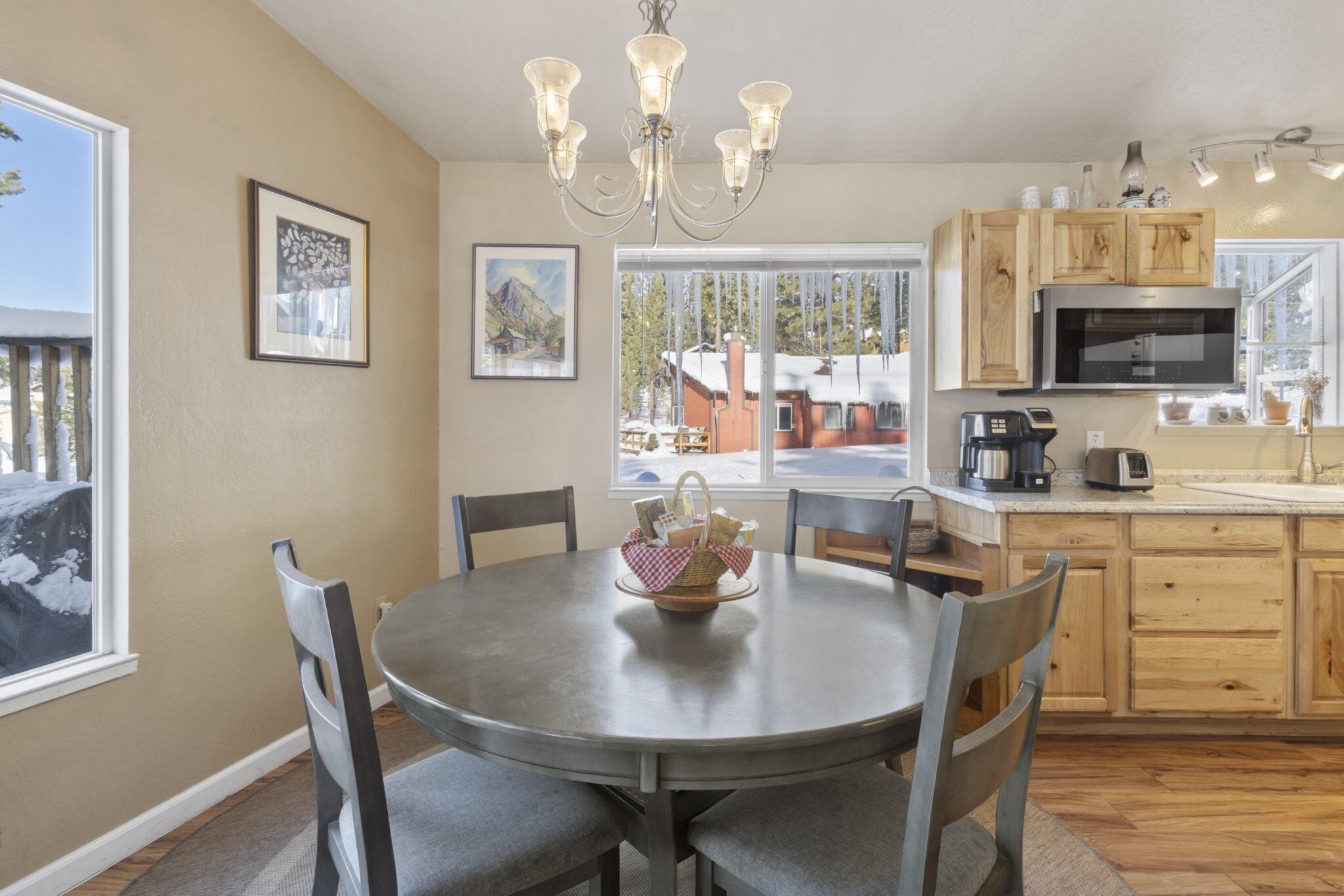 Una cocina con una mesa de comedor redonda, sillas, una araña, armarios de madera, un microondas y una ventana con vista a una casa.