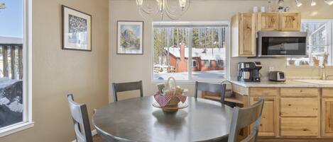 Una cocina con una mesa de comedor redonda, sillas, una araña, armarios de madera, un microondas y una ventana con vista a una casa.