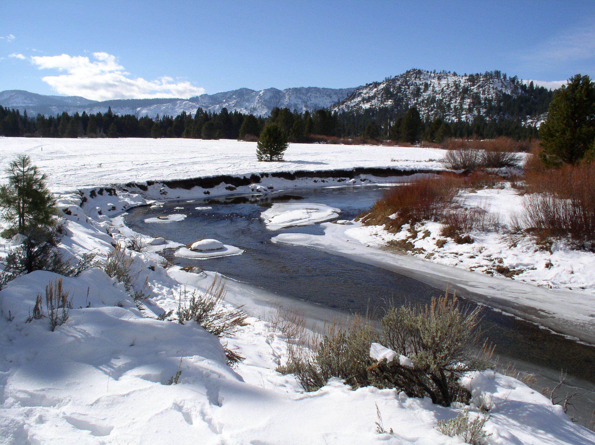 A snow-covered landscape with a river, mountains, and sparse vegetation.