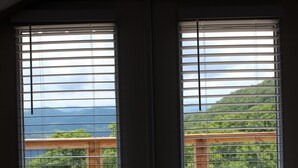 A room with two windows with horizontal blinds, a wooden deck outside, and a view of a mountainous landscape.